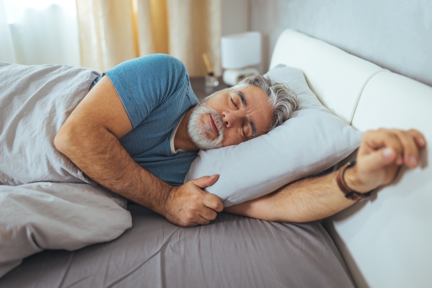 A man with grey hair and a grey mustache sleeps in bed.