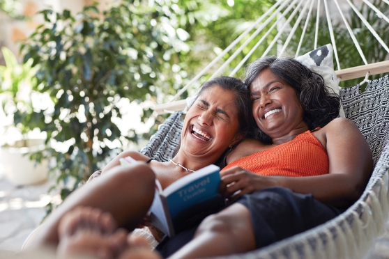 Two women laugh while laying in a hammock together and reading a book