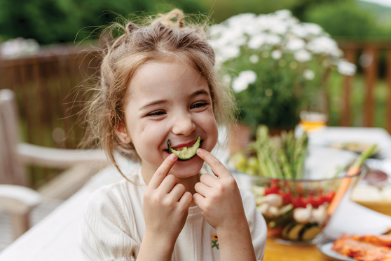 A little girl holds a cucumber coin (with a bite out of it) up to her lips, as if it's a smile.