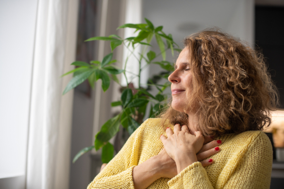 A woman sits with her hands across her chest, wearing a yellow sweater and a serene expression on her face.