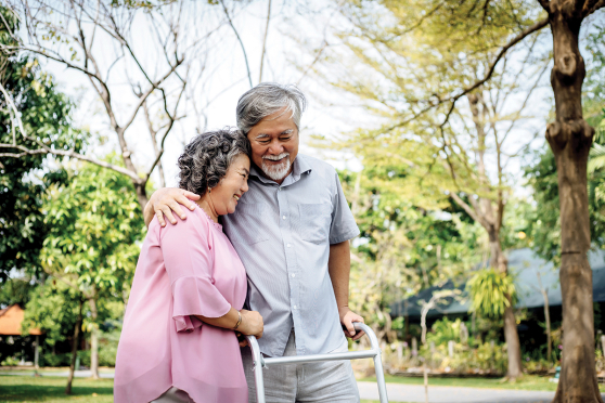 An older couple walks together. The man uses a walker and has his arm affectionately around the woman.